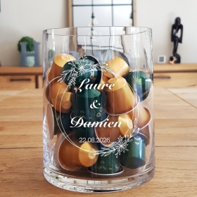 Un vase en verre avec une couronne de fleurs, gravé, pour de futurs mariés, avec leur prénom et date de leur mariage.